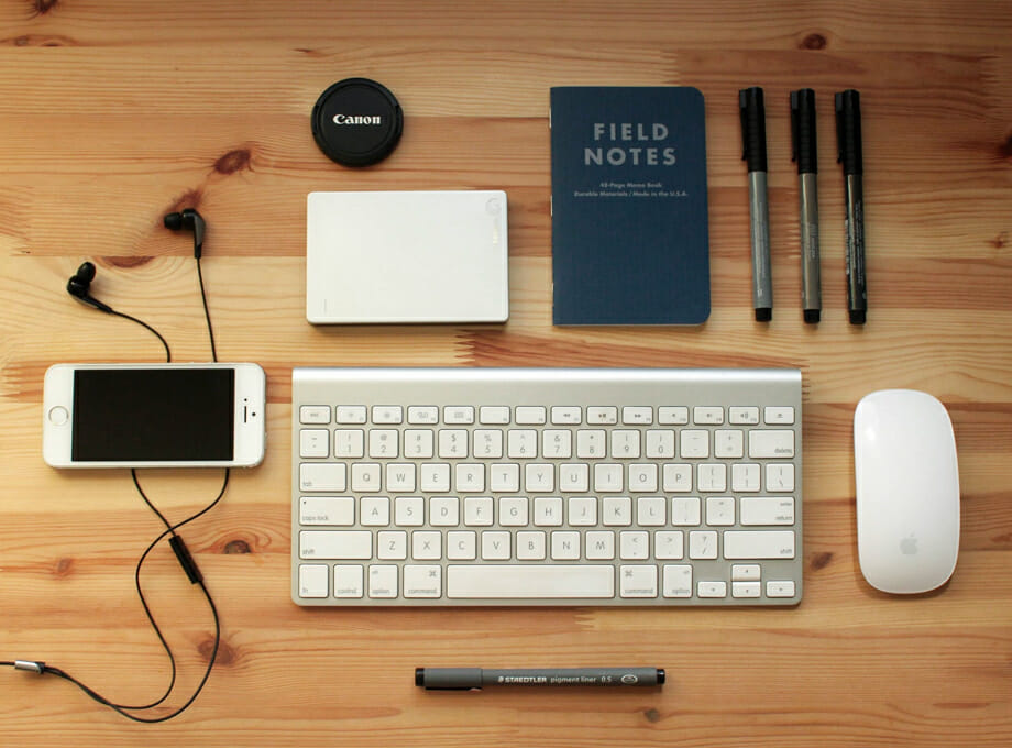 A staged photo of a keyboard, mouse, mobile phone, pens, and notebook.
