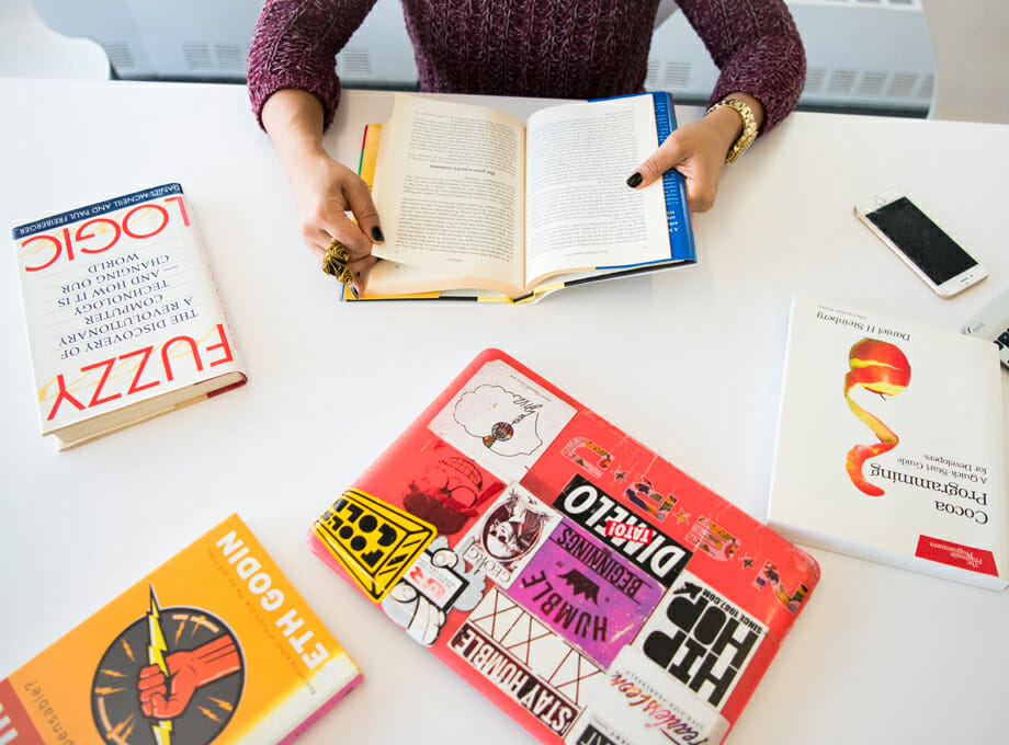 A person holds an open book at a desk cluttered with more books, a phone, and a laptop.