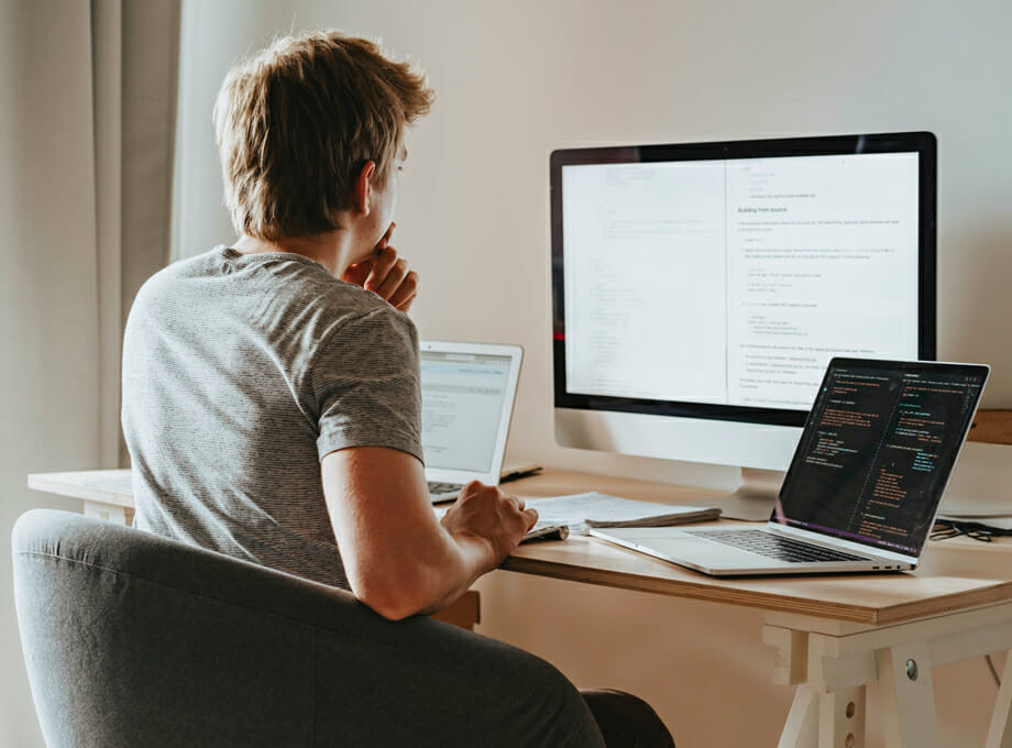 A young adult sitting at a desk concentrates on a large monitor with two laptops nearby.