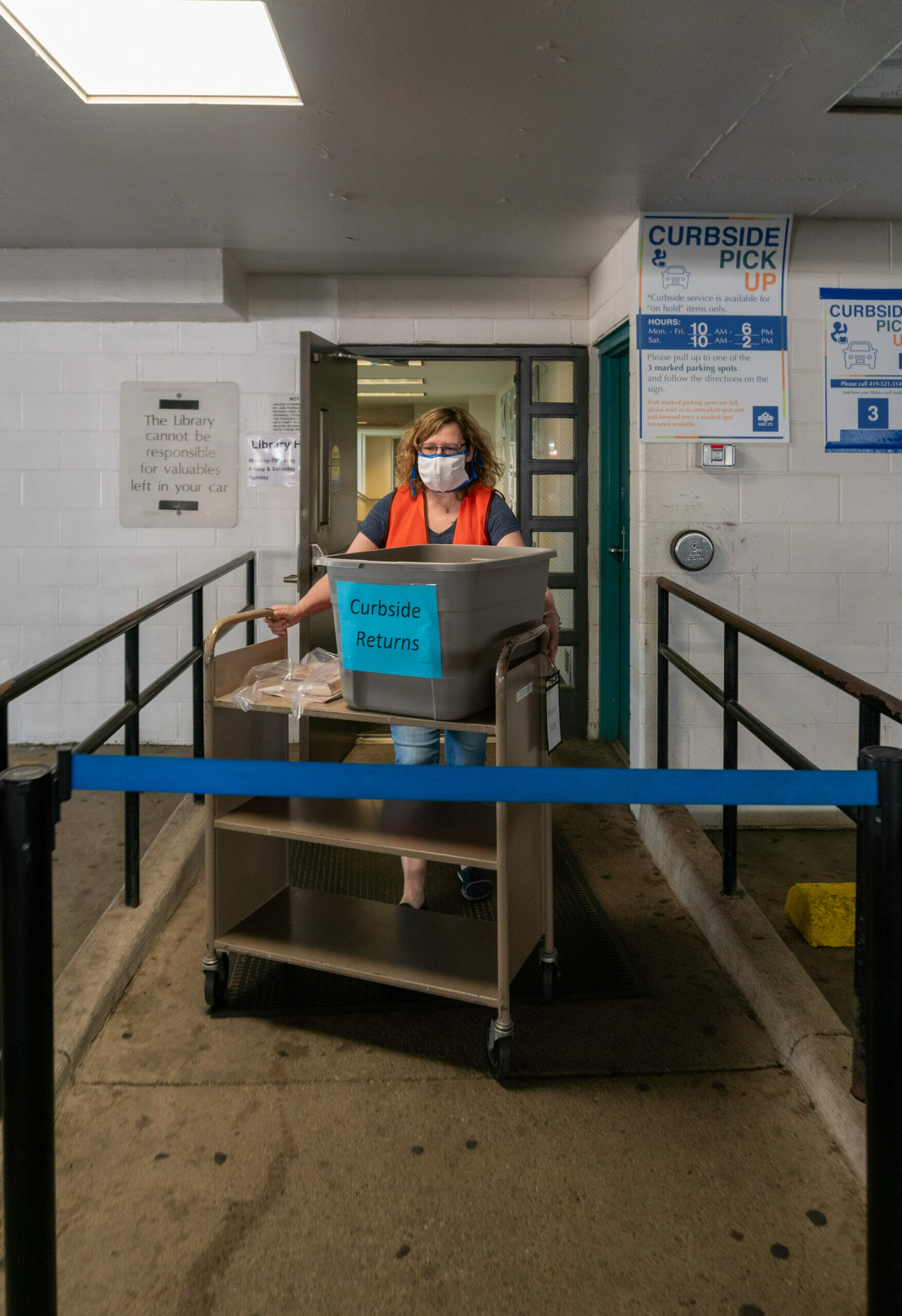 A masked staff member wheels a cart of curbside pick up materials out to a car in the parking garage at the Main Library.