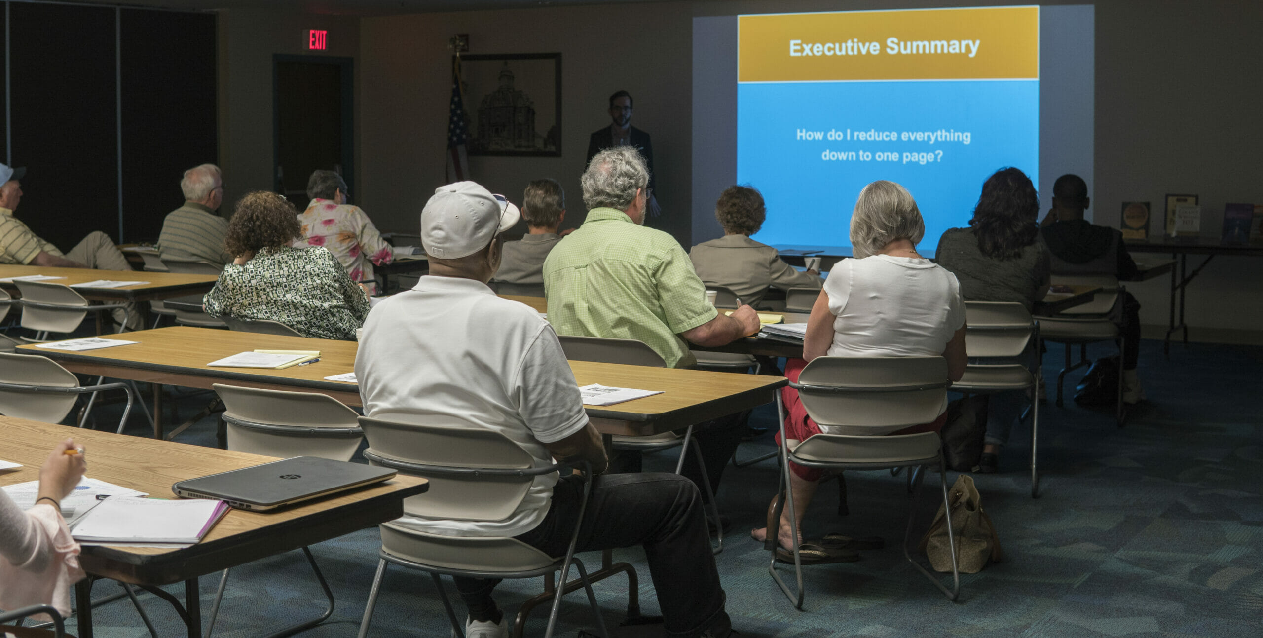 A group of adults seated at classroom style seating listening to a speaker with a presentation in the Community Room at the Main Library