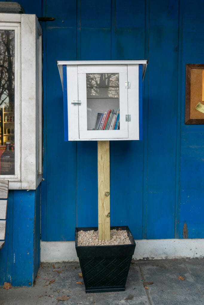 A white, MRCPL little free library box stands outdoors in front of a blue wall.