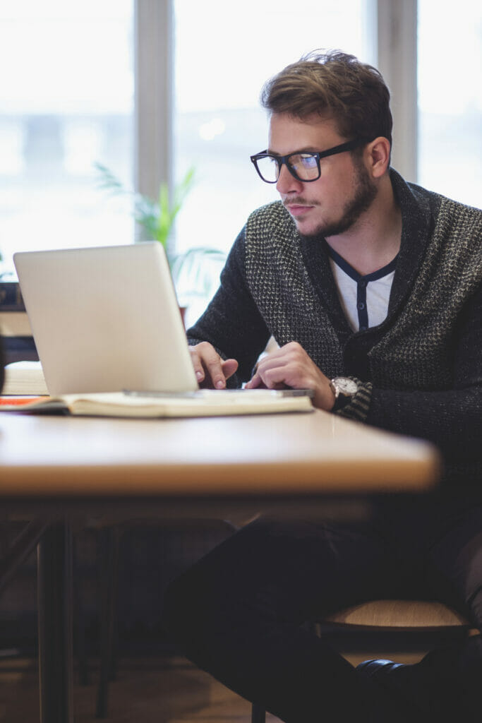 A person sits at a table near a window and studies the screen of a laptop while using the keyboard.