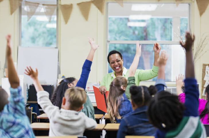 An African-American teacher, a mature woman in her 40s, sitting in front of her class of elementary school students, 6 and 7 years old, reading a book. She has asked a question and most of the children are raising their hands. They are in first grade or second grade.