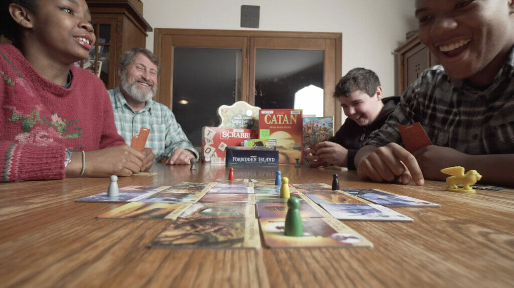 A family of three teenagers and a father play a board game seated at a table in the family's home.