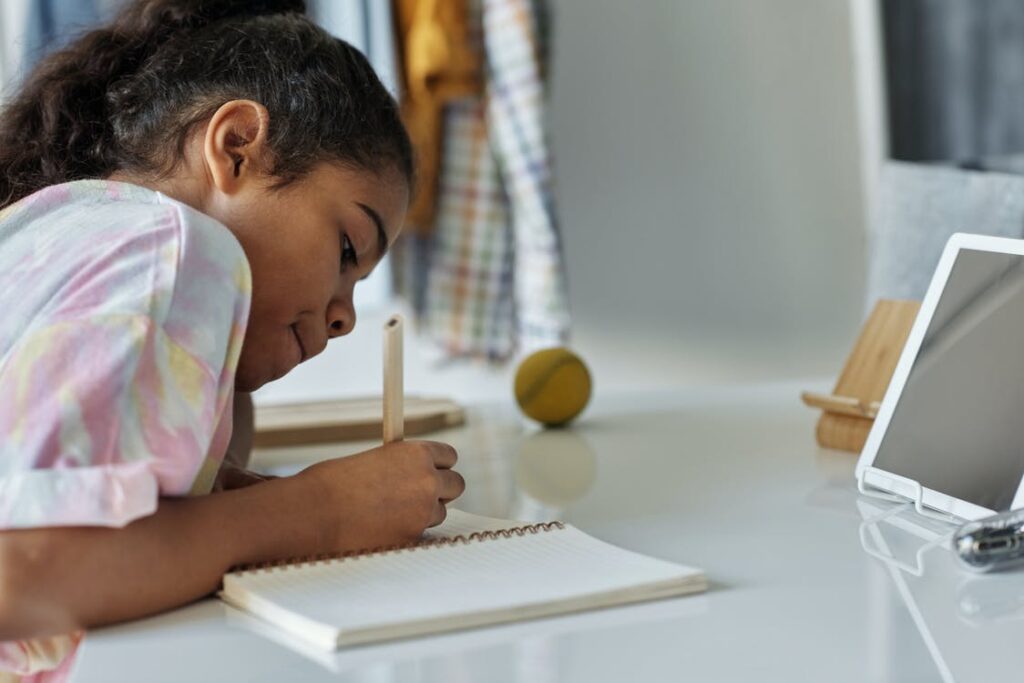 A child concentrates on writing in a notebook seated at a table in front of an electric tablet.
