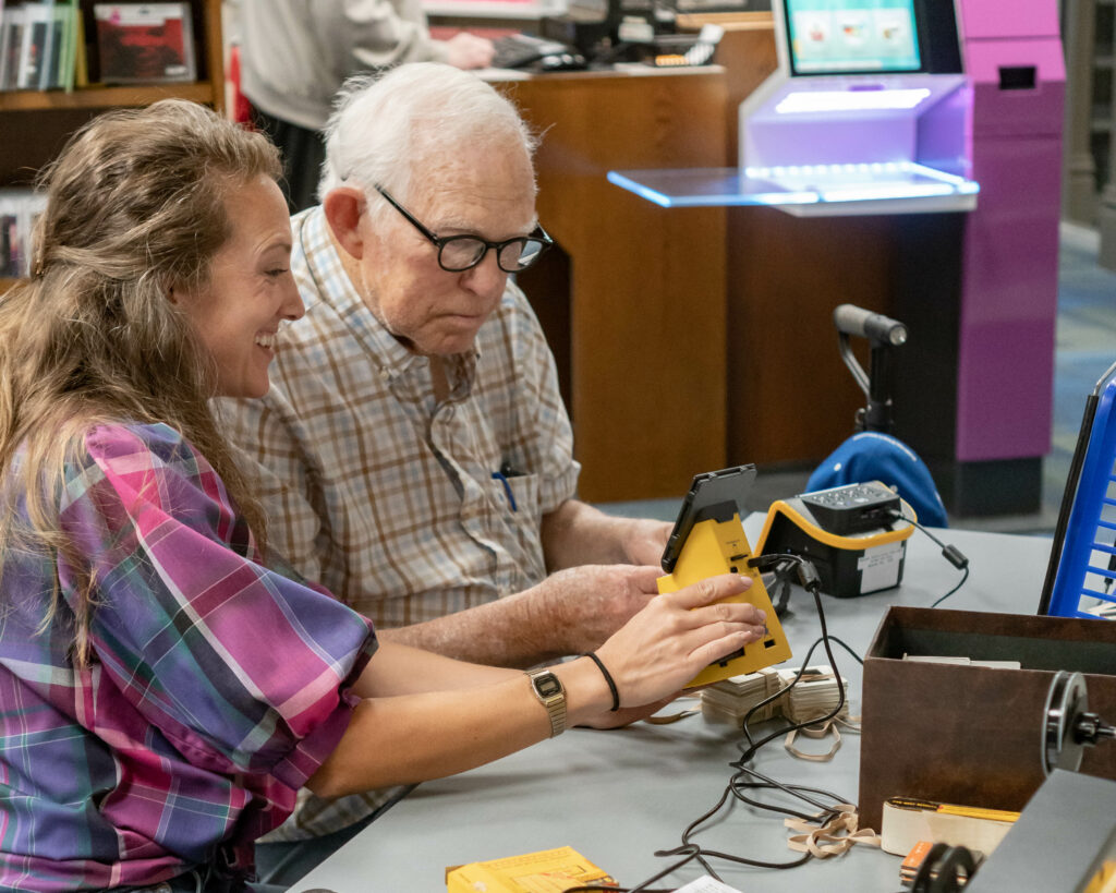 A person happily explains technology to an elderly person while seated at the Main Library Memory Lab.