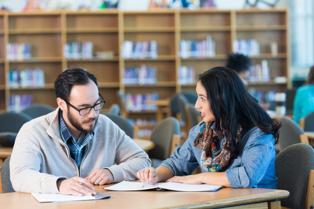 2 people sitting at table in library writing notes and studying. To people blurred in background reading at table.