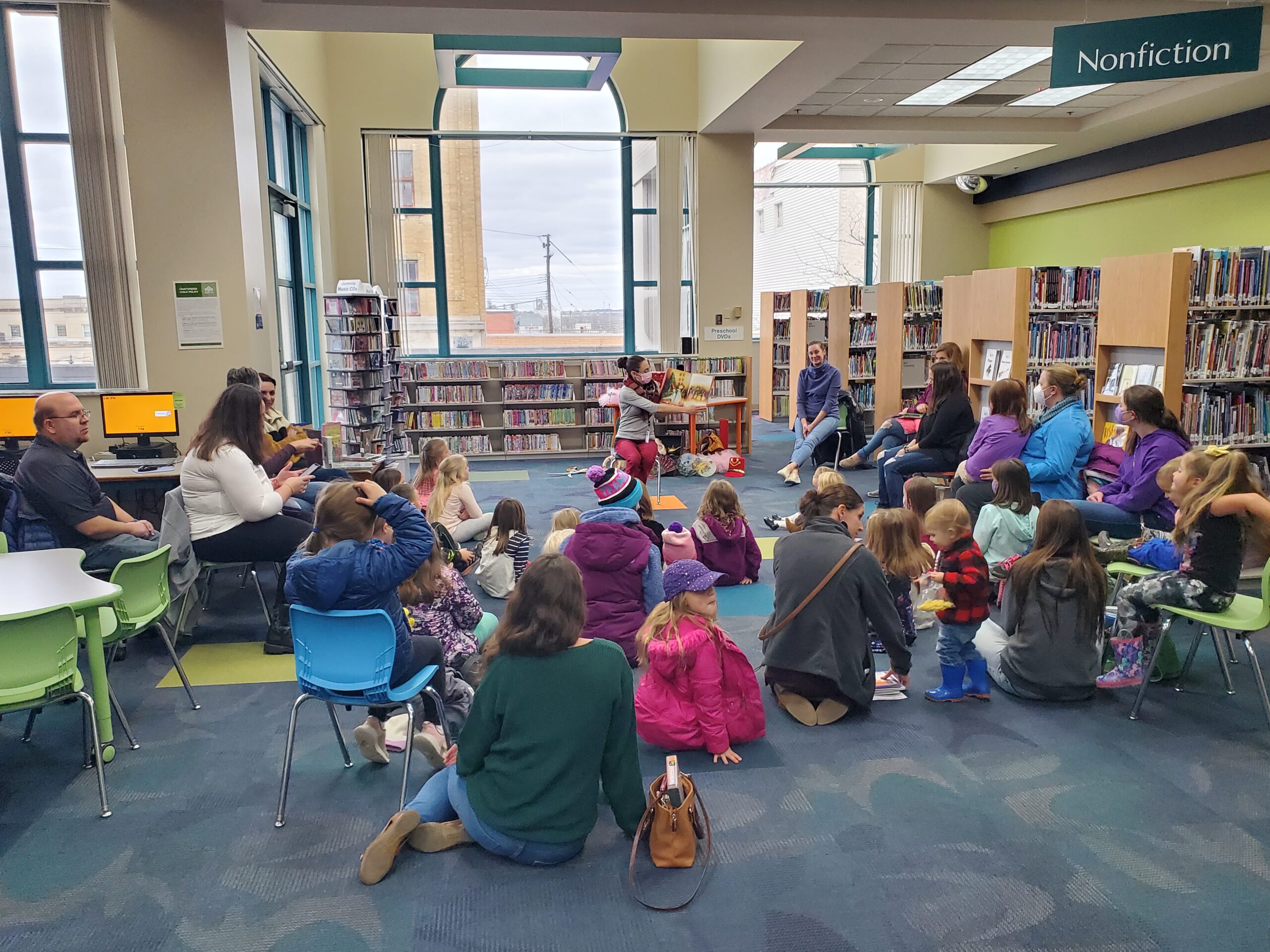 Children and adults sitting on chairs and on the floor listening to librarian read a story for story time.