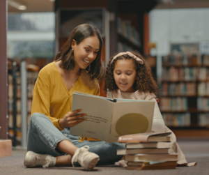 Adult and child sitting on floor reading a book together. A pile of books in front of them on floor. Blurry background image of books on shelves.