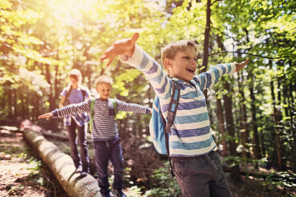 3 young children balancing on a log while hiking in the woods with backpacks on a sunny day.