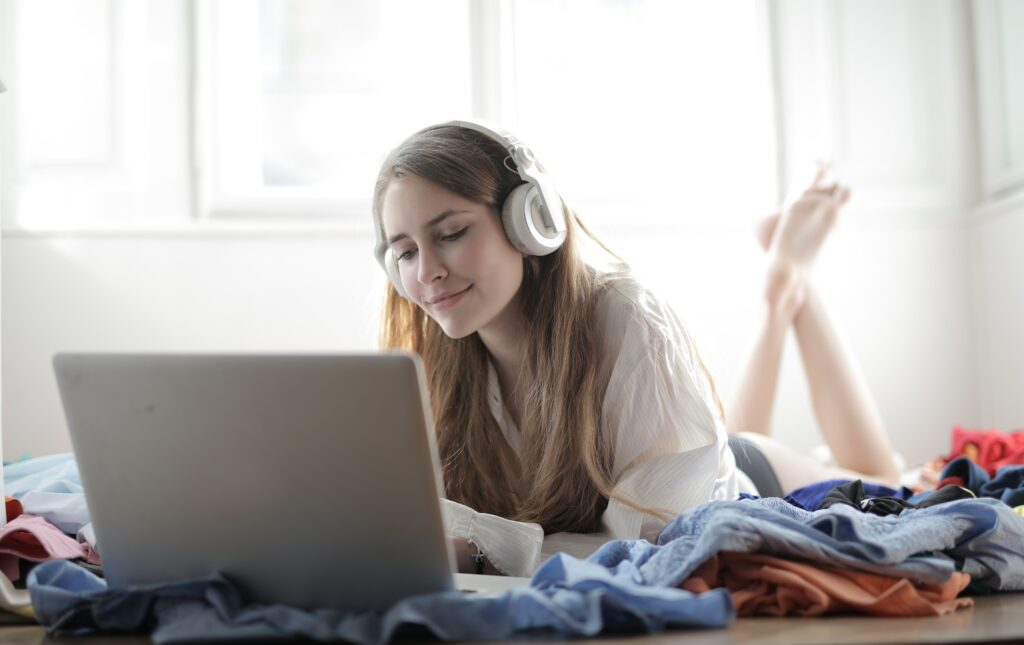 person laying on stomach with headphones on looking at laptop and clothes scattered around her on the floor.