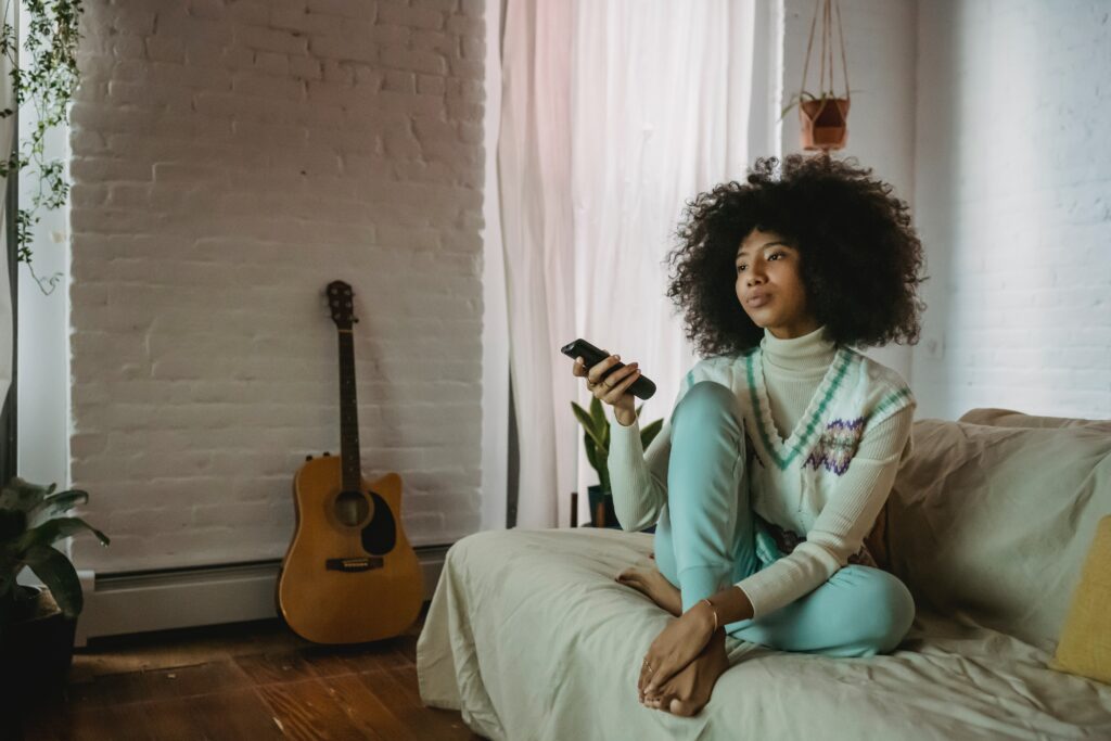 Adult sitting on couch with tv remote. Hanging plant behind her and guitar leaning against a white brick wall
