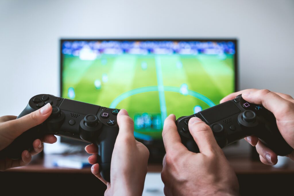 Close-up of two individuals hands holding video controllers playing sports game on blurred image of a TV.