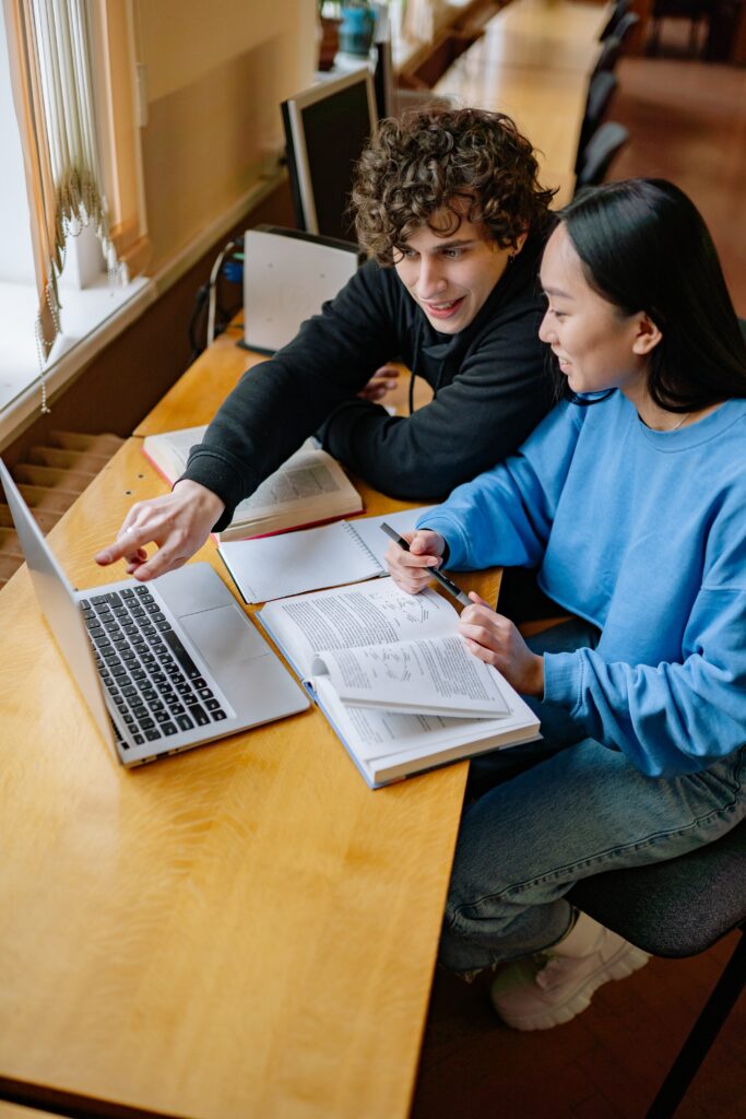 Two adults sitting side by side at a table with textbooks and a laptop