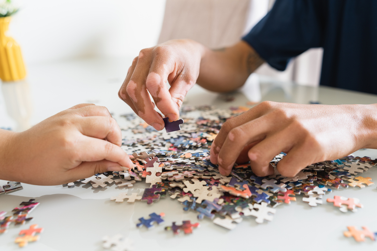 Detail of hands of two individuals looking for puzzle pieces on table.