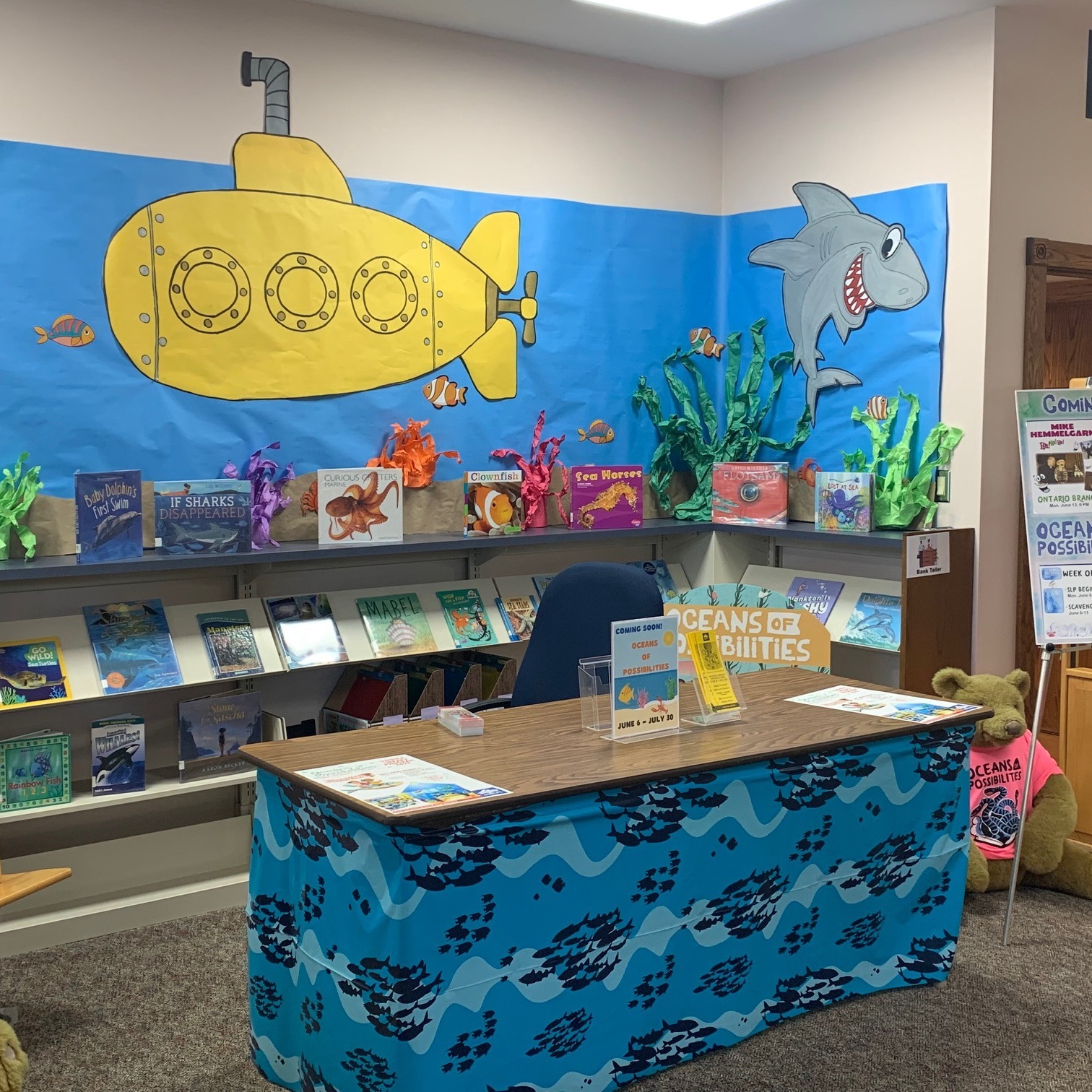 oceans of possibilities summer library program display. books on shelves. pictures of submarine, shark, fish, water made from colored paper on wall above books