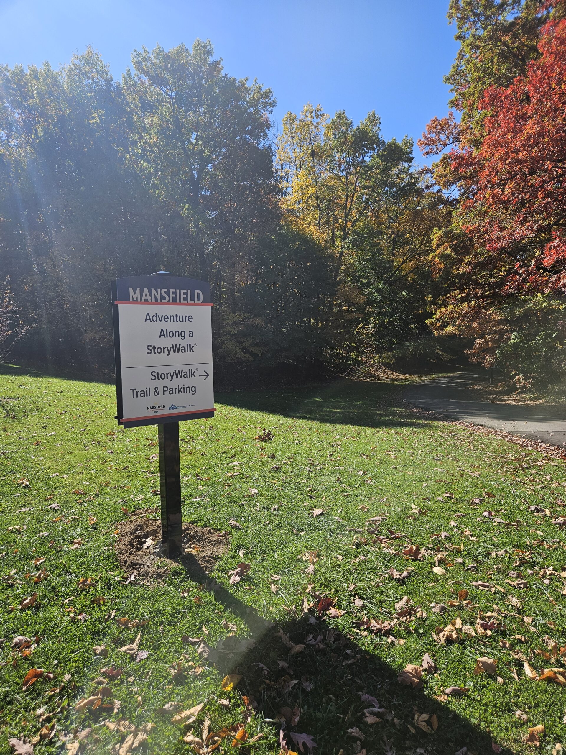 White Mansfield City Park sign on a sunny day with fall foliage in the background