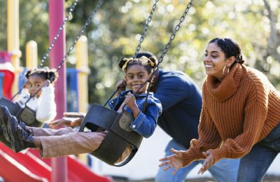 A young family playing on a playground. The children, two daughters, are sitting on swings with the parents standing behind them. The focus is the mother and older girl, 5 years old, in the foreground. The child is smiling at the camera. The father and 3 year old daughter are out of focus in the background. The children are mixed race Hispanic, African-American and Native American.