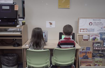 Two children play on tablets seated at a table in the Library.